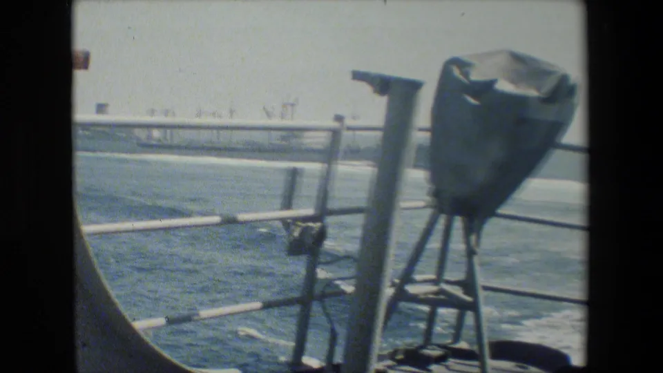 The camera looks from the ship deck past railings and a mounted covered object toward open water and distant industrial or port structures.