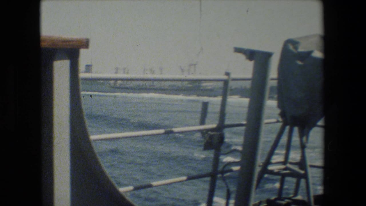 Color-faded 8mm frame showing a ship deck railing, open water, wake, and distant industrial-looking shoreline structures from a naval vessel context identified by the clip metadata.