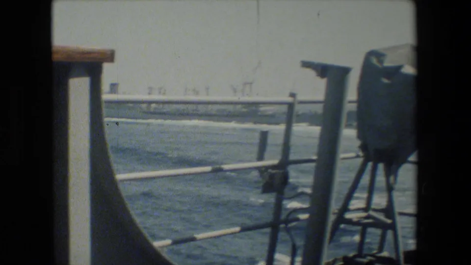 Color-faded 8mm frame showing a ship deck railing, open water, wake, and distant industrial-looking shoreline structures from a naval vessel context identified by the clip metadata.