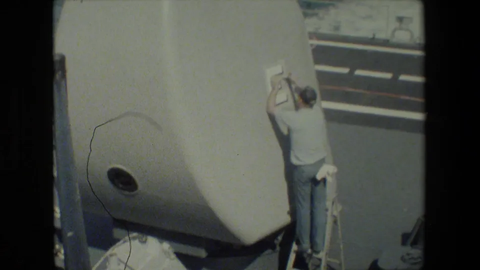 A crew member in a white shirt and cap stands beside a large white gun turret on a ship deck, with railings and ocean visible in the background.