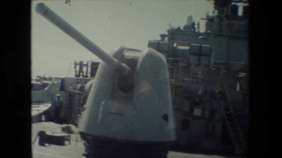 Shipboard view of a white gun turret and nearby deck equipment, with parts of the vessel structure and open water visible in grainy 8mm footage.
