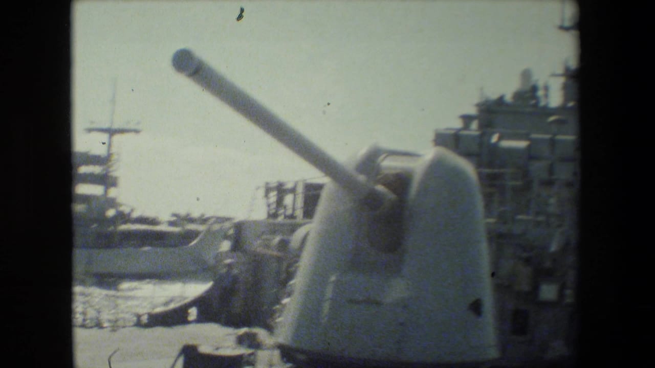 Naval gun turret in the foreground with a docked warship and masts behind it.