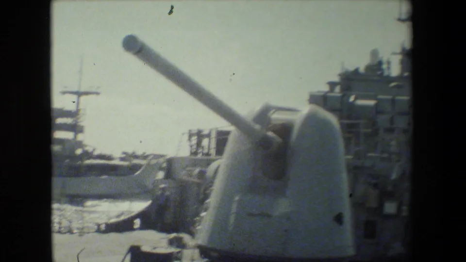 Soft 8mm frame of a naval deck gun turret in the foreground, with another docked vessel, superstructure, masts, and harbor water visible behind it.