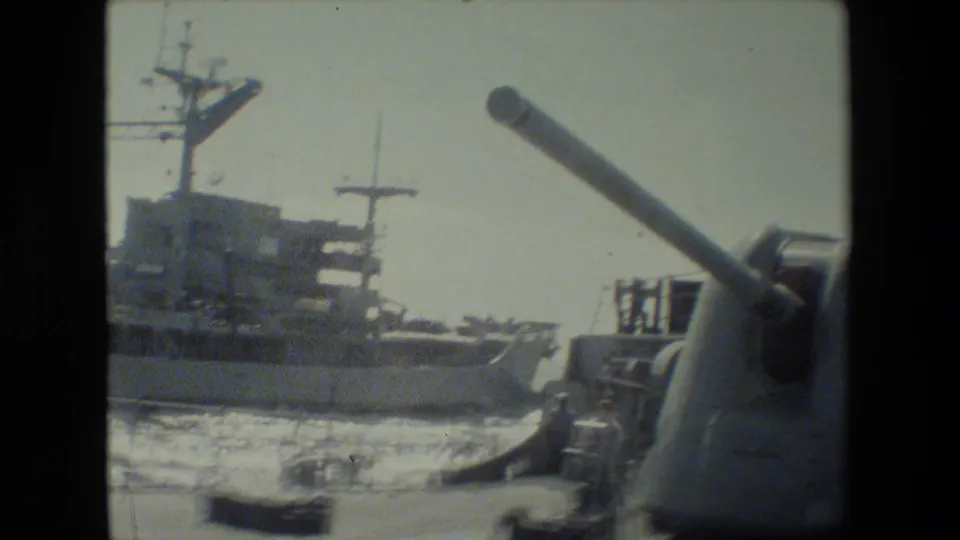 The camera frames the gun turret at the right edge while a nearby naval vessel, mast structures, and bright harbor water appear across the background.