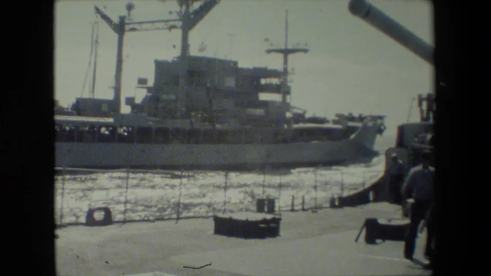 A docked naval vessel is seen broadside across the harbor, with its superstructure and mast visible and part of the deck gun barrel cutting through the upper right of the frame.