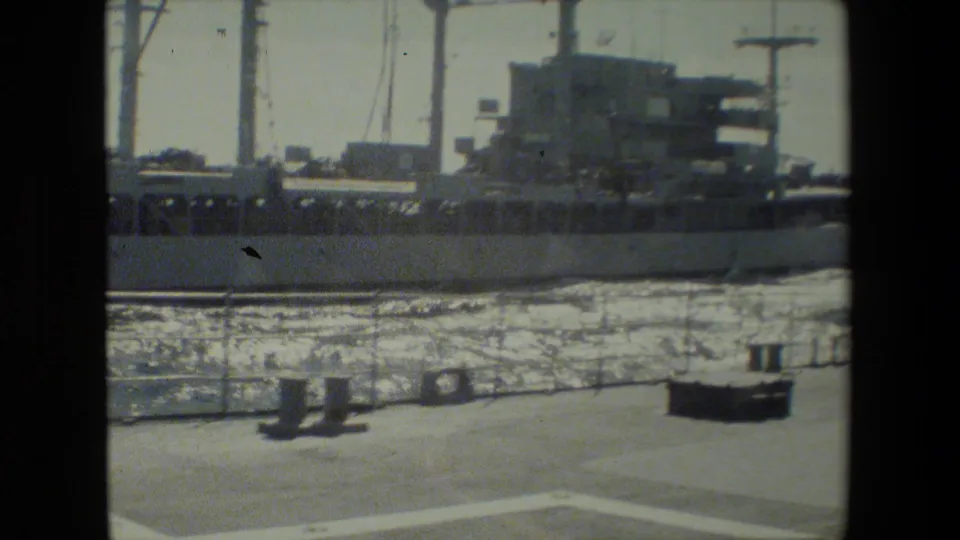 The frame shows a docked naval vessel across rippled harbor water, with dockside posts and equipment in front and a small human figure near the right edge.