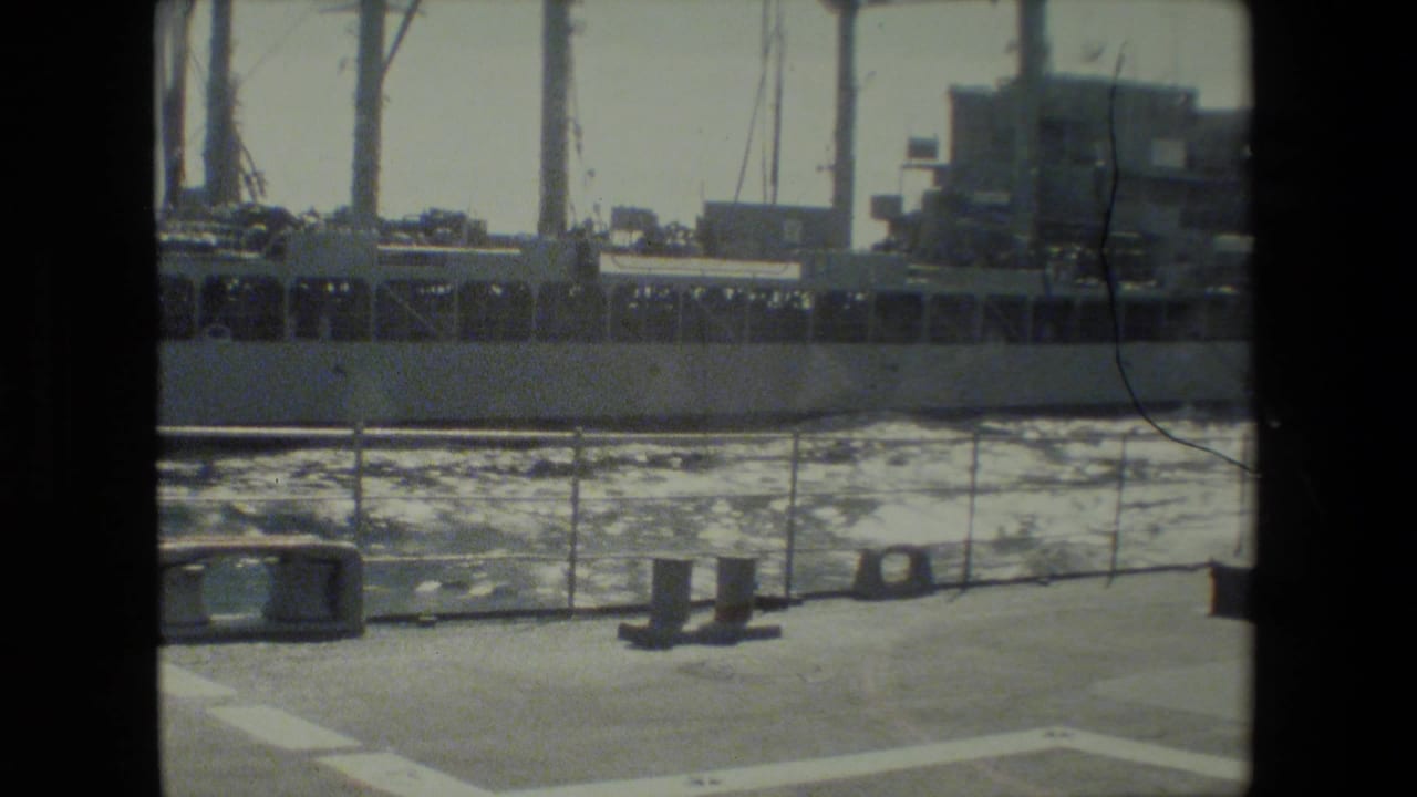 Docked ship seen across harbor water from a deck with railings and fittings.