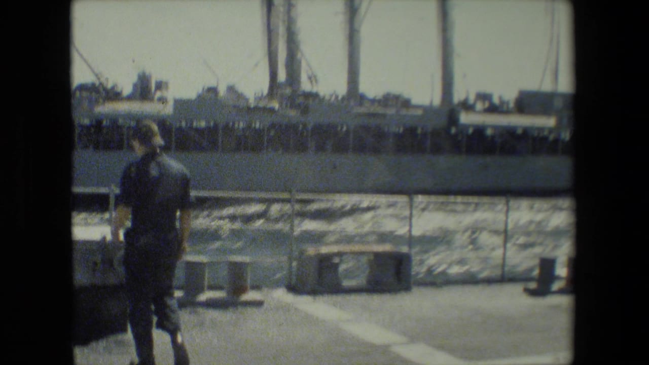 Uniformed sailor walking on deck near rail with docked ship behind him.