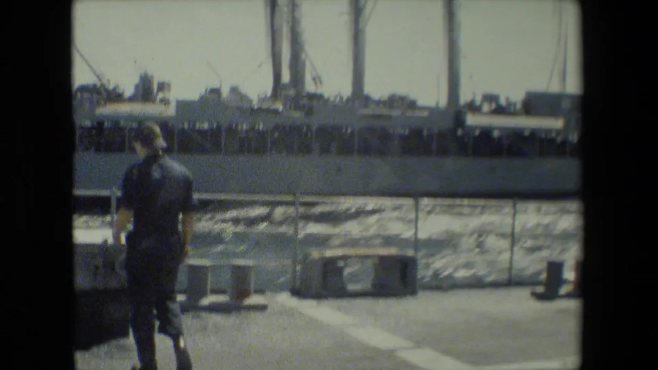 A uniformed adult sailor walks along the deck near a railing, with moving water and a large docked ship in the background.