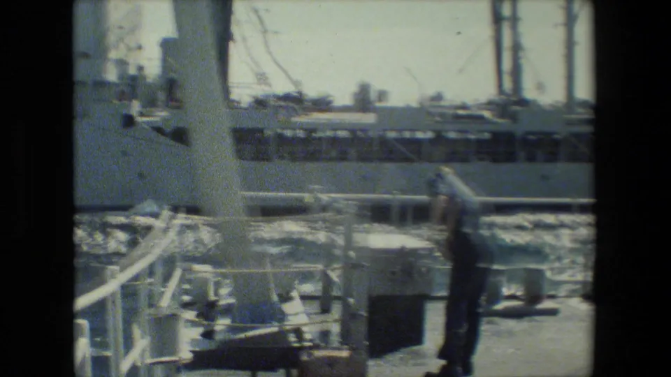 A handheld 8mm frame catches a sailor near deck equipment and railings, with choppy water and the side of a docked ship behind him.