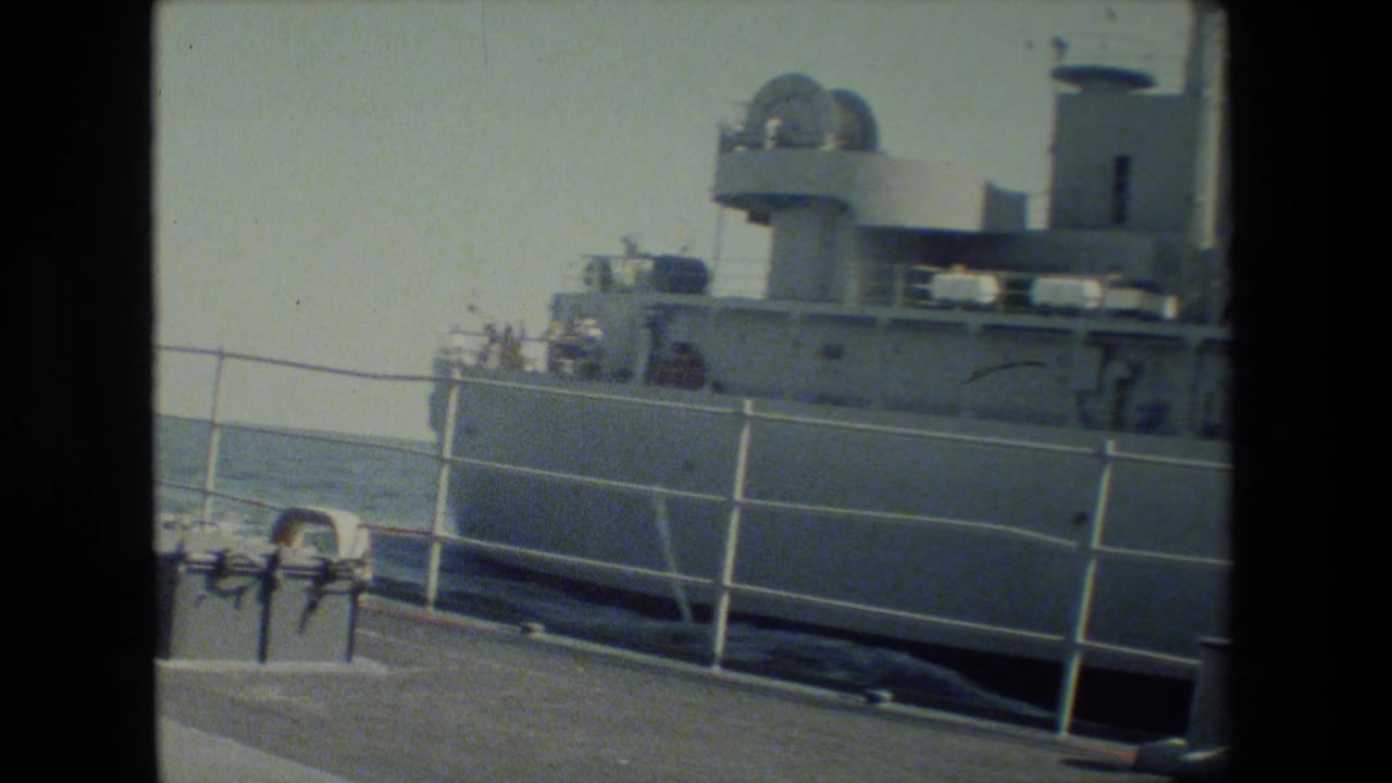 A gray docked research or naval vessel appears beside a pier, with white railings, deck equipment, open water, and a small person in a red shirt near the upper railing.