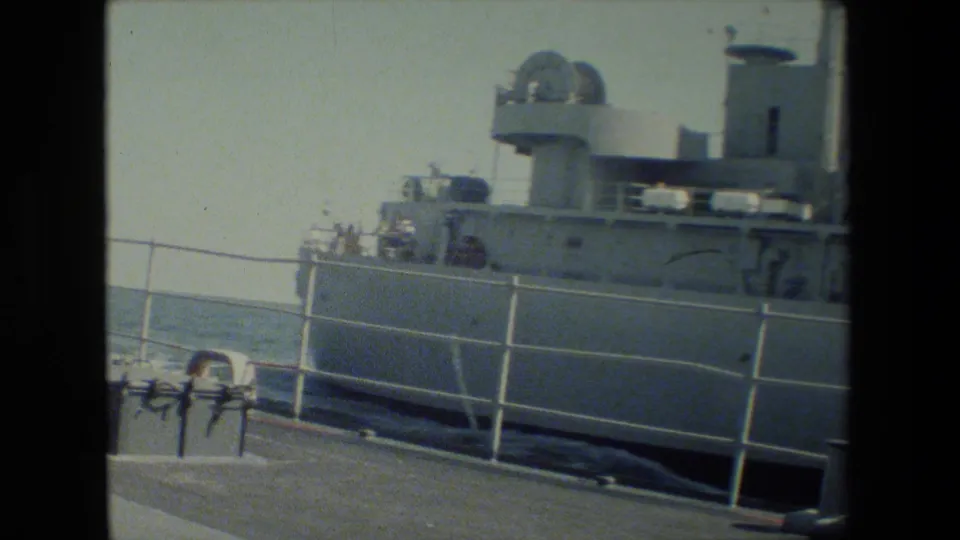 A gray docked research or naval vessel appears beside a pier, with white railings, deck equipment, open water, and a small person in a red shirt near the upper railing.