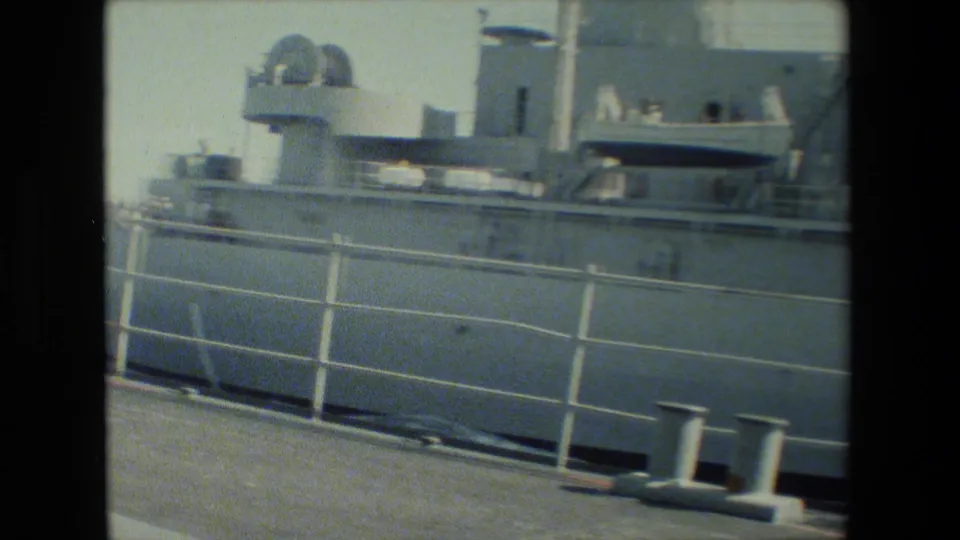Side view of the gray docked vessel from the pier, showing the railings, deck structures, hull side, bollards, and sunlit dock surface in soft 8mm color.