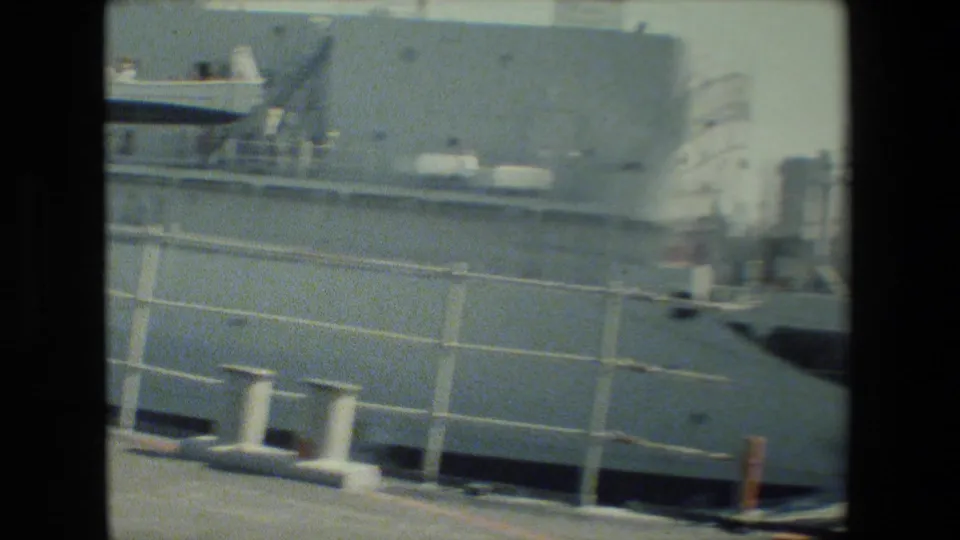 A closer, slightly blurred 8mm view of the vessel side shows white railings, gray deck structure, mooring bollards, and the pier edge under bright daylight.