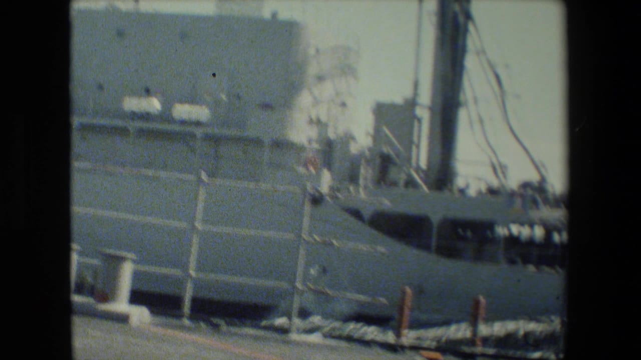Soft 8mm frame showing part of a gray U.S. Navy warship near dockside equipment, with harbor structures in the background and no people clearly visible.