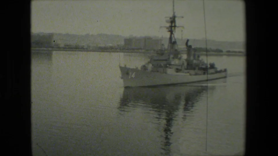A gray naval warship appears underway or at slow harbor speed, with its mast reflected in calm water and distant shoreline buildings behind it.