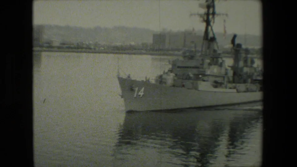 The bow and forward section of a gray naval ship marked 14 are visible in soft 8mm footage, with calm water and a hazy shoreline in the background.