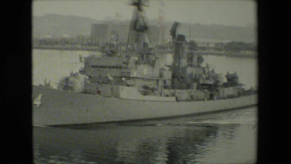 Side view of the gray naval vessel shows its mast, antennas, deck equipment, and partial bow number as it sits or moves slowly in harbor water.