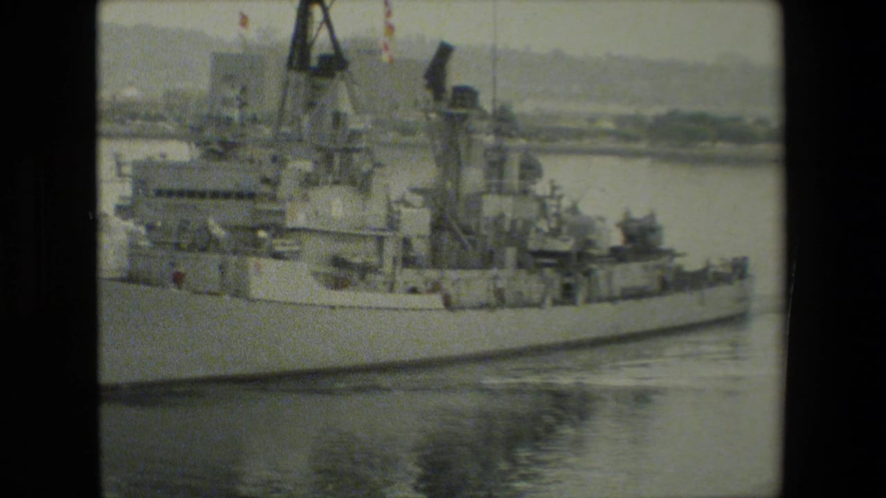 Grainy 8mm frame of a gray U.S. Navy ship viewed broadside in calm harbor water, with deck equipment and industrial structures visible behind the vessel.