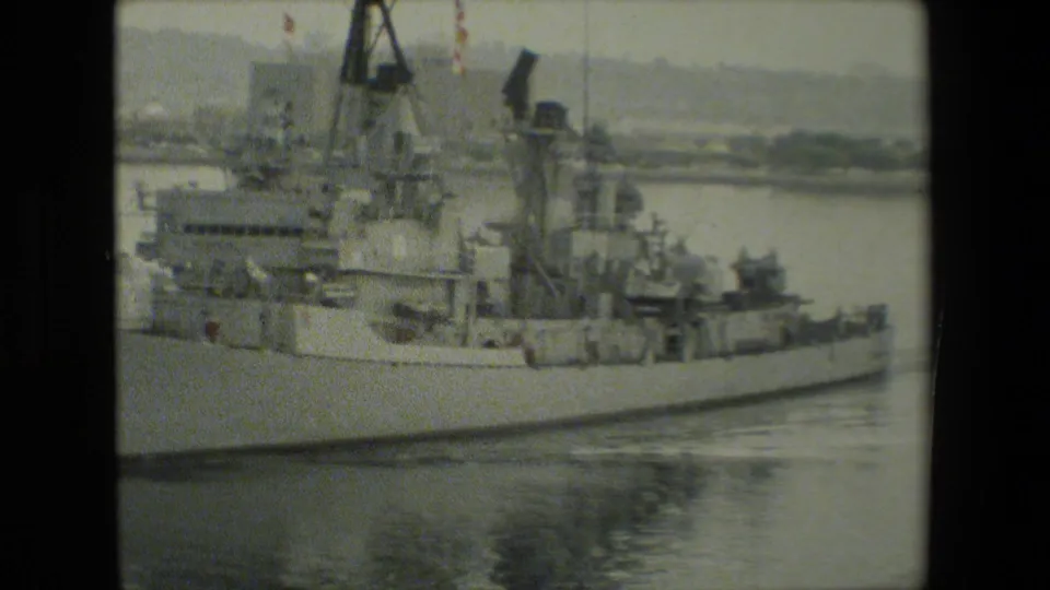 Grainy 8mm frame of a gray U.S. Navy ship viewed broadside in calm harbor water, with deck equipment and industrial structures visible behind the vessel.