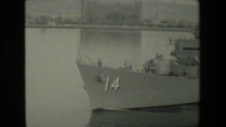 Close 8mm view of a gray naval ship's bow bearing the number 14, with a partial view of deck structures and calm harbor water beside the hull.