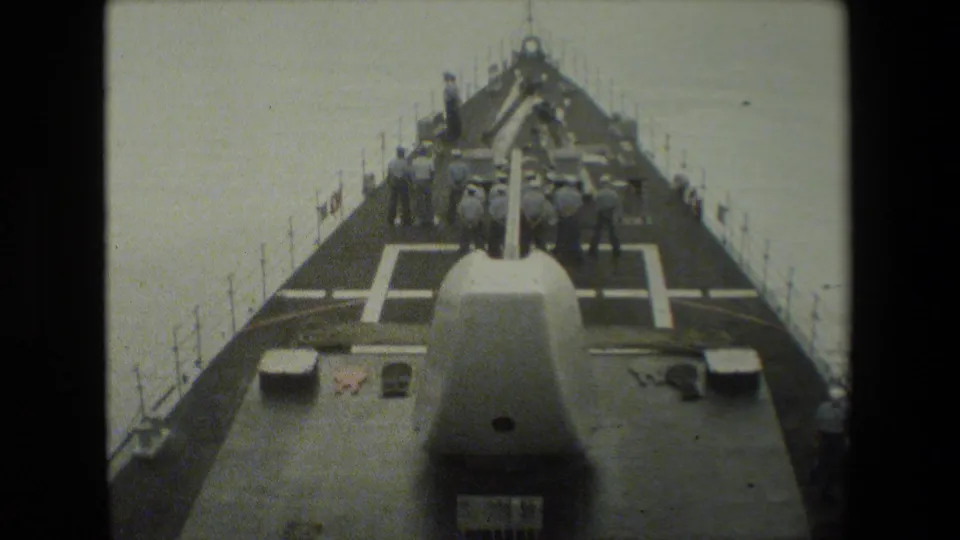 View looking along the forward deck of a naval vessel, with deck guns or equipment, railings, and several crew members visible near the bow area.