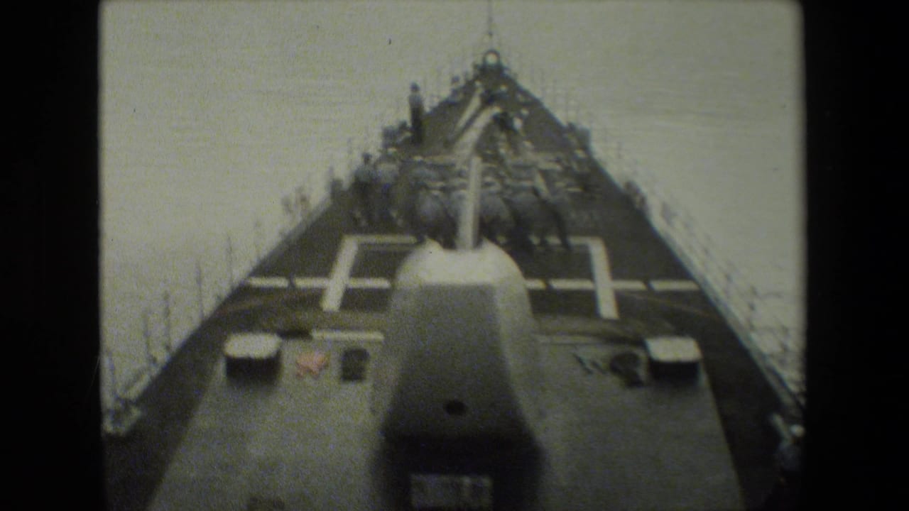 Forward-looking 8mm frame from a naval vessel shows uniformed crewmen on deck near a large gun turret as the ship moves over calm water.