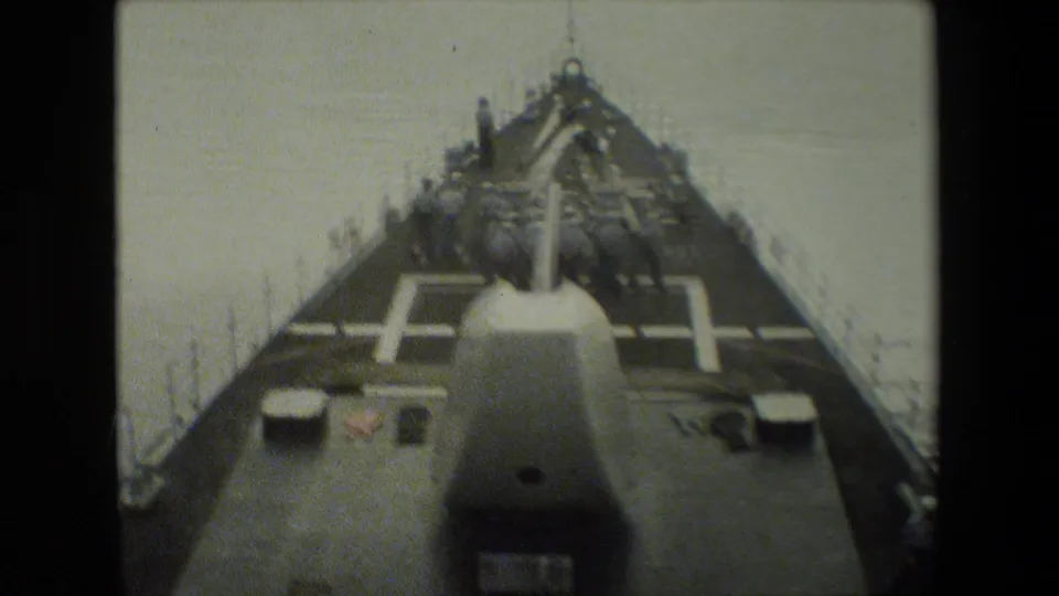 Forward-looking 8mm frame from a naval vessel shows uniformed crewmen on deck near a large gun turret as the ship moves over calm water.