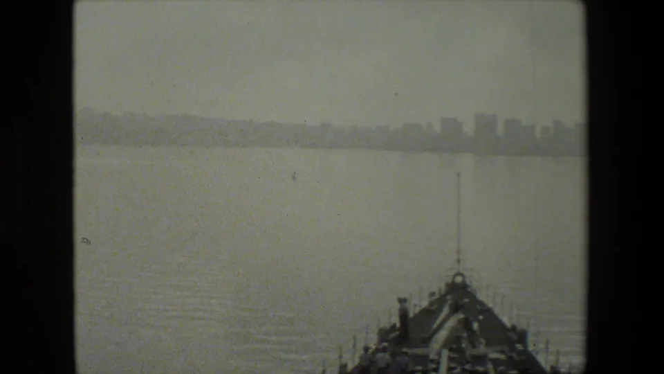 A hazy waterfront skyline appears ahead of the ship, with the vessel's bow and deck crew partially visible at the bottom of the 8mm frame.