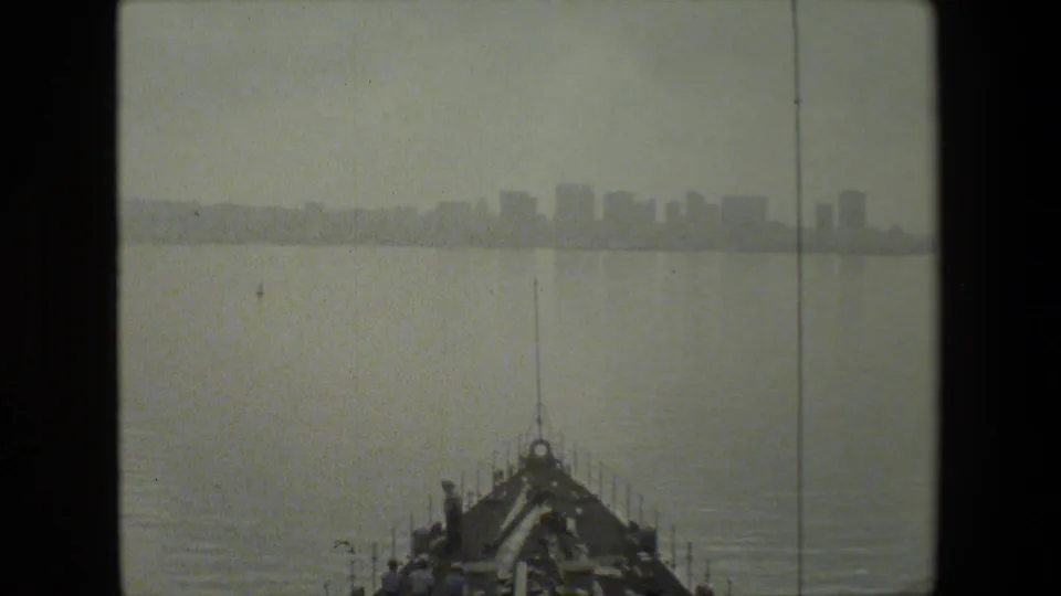 From the bow of the naval vessel, the frame shows a hazy city skyline across the water with crewmen and deck hardware visible in the foreground.