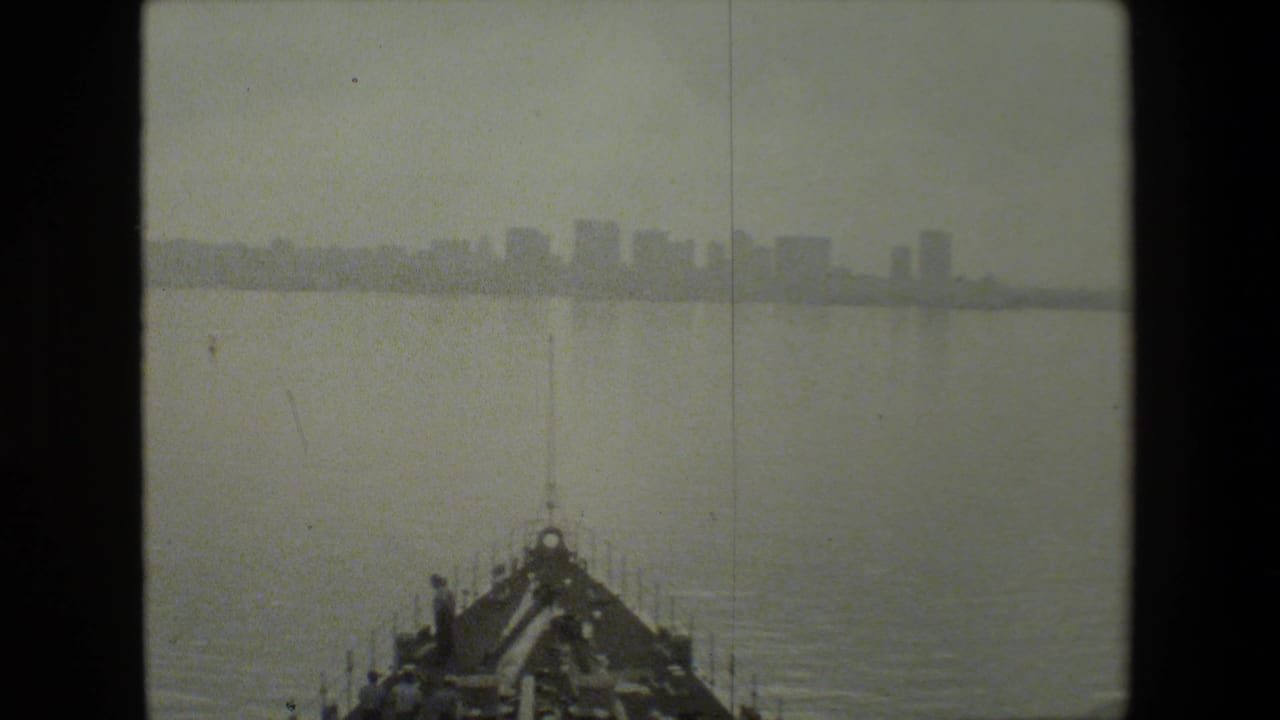 Black-and-white 8mm frame showing the bow of a vessel moving through calm harbor water with a hazy urban skyline in the distance.