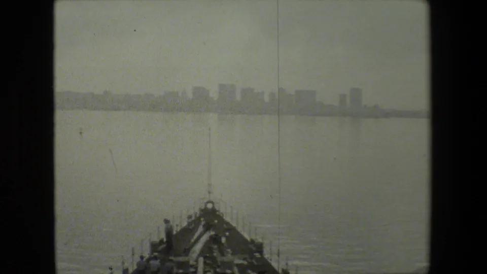 Black-and-white 8mm frame showing the bow of a vessel moving through calm harbor water with a hazy urban skyline in the distance.
