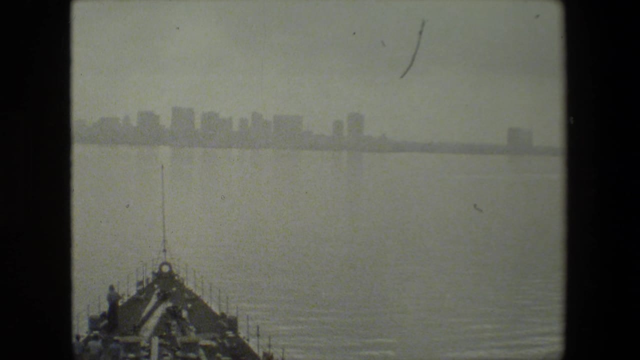 Ship bow moves through water with a distant city skyline beyond it.