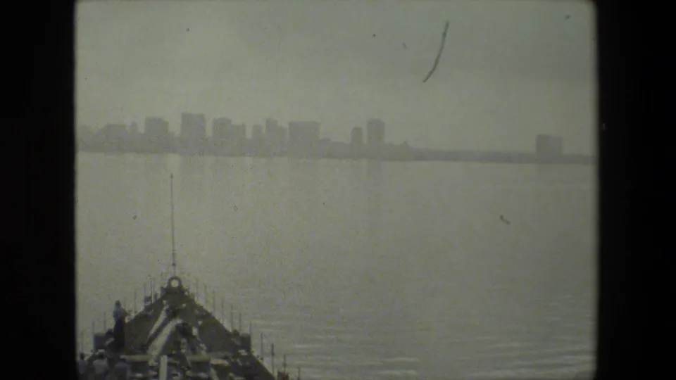 Black-and-white home-movie frame of a vessel bow cutting through harbor water, with a soft distant skyline and film gate borders visible.