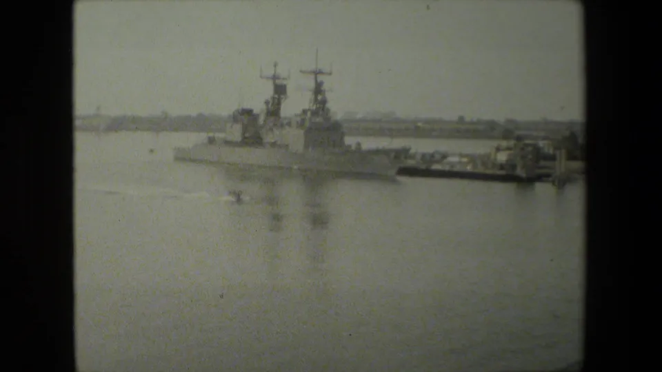 Black-and-white 8mm frame showing two large naval vessels near a pier or dock, with calm harbor water and shoreline haze behind them.