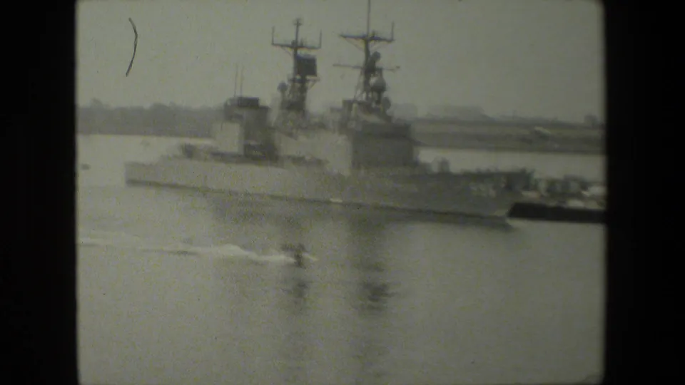 Black-and-white home-movie frame of a large naval vessel beside harbor structures, with a smaller boat wake crossing the foreground.