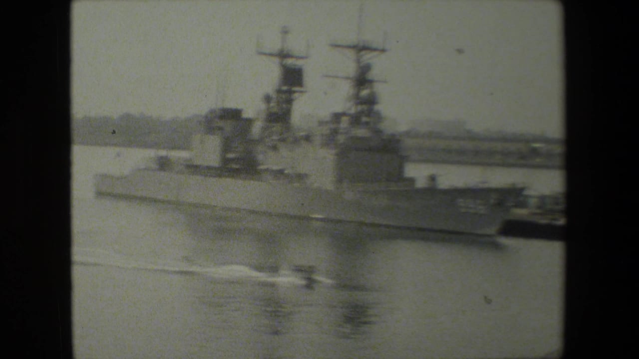 Grainy 8mm frame of a large U.S. Navy warship at dock, viewed from across calm water in footage supplied as 1981 naval-base material.