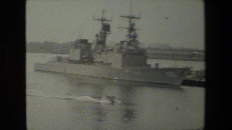 Grainy 8mm frame of a large U.S. Navy warship at dock, viewed from across calm water in footage supplied as 1981 naval-base material.