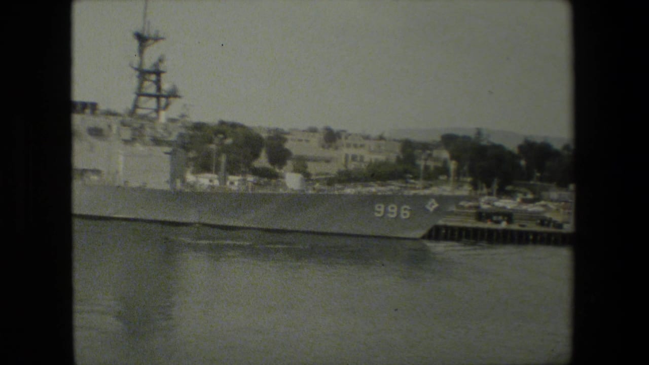 Bow and side of a naval vessel marked 996 beside a dock, with shoreline buildings behind it.