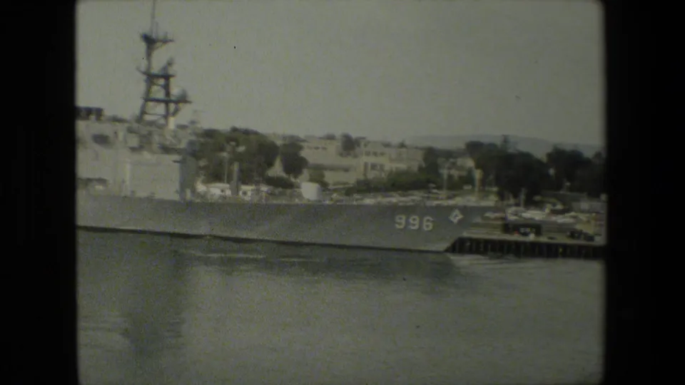 Naval vessel with visible hull number 996 rests beside a dock, with masts, deck equipment, harbor water, and low shoreline buildings in the background.