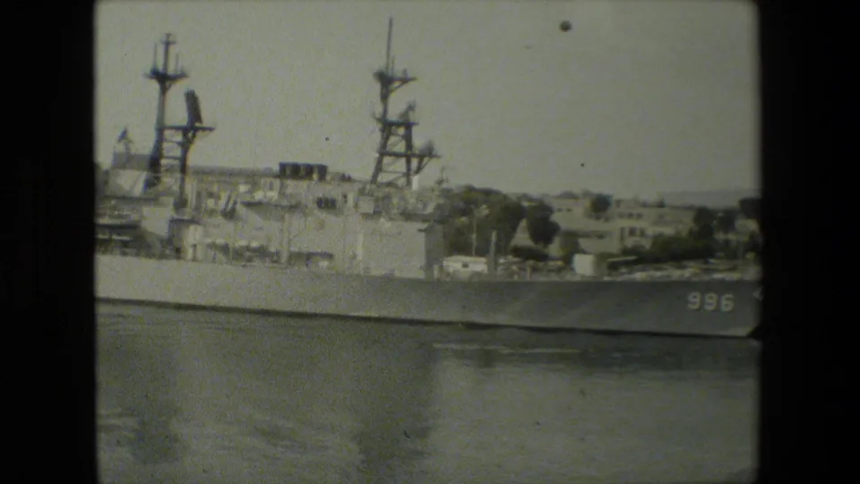 Closer grainy view of the docked naval vessel shows hull number 996, mast structures, upper decks, and shoreline buildings beyond the harbor.