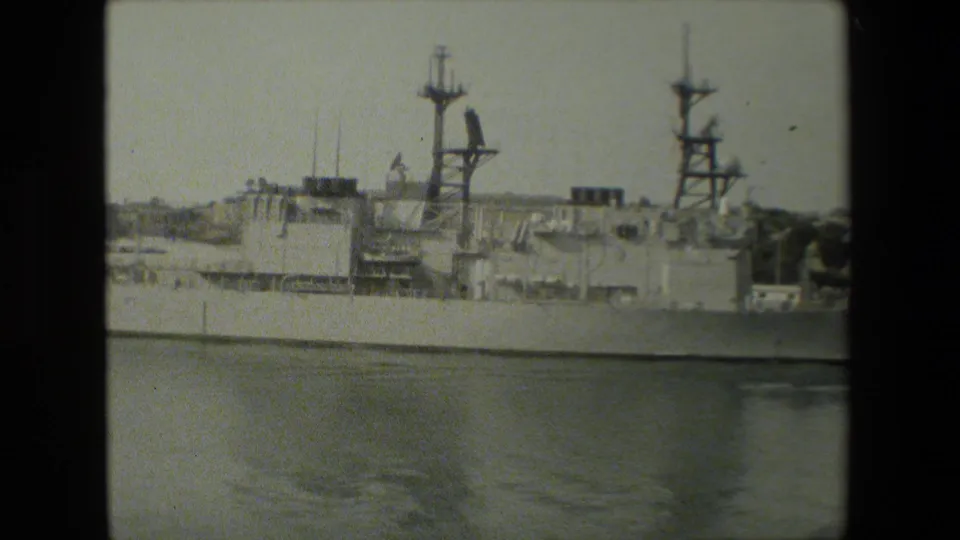 Side-on 8mm frame of the docked warship shows its superstructure, two prominent mast assemblies, calm water reflections, and harbor shoreline in the distance.