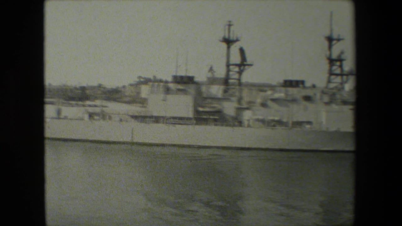 A large docked naval vessel fills the frame in grainy black-and-white 8mm footage, with superstructure, mast shapes, and calm harbor water visible in front.