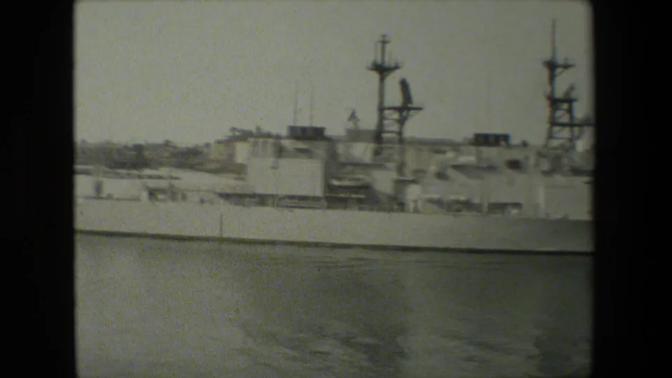 A large docked naval vessel fills the frame in grainy black-and-white 8mm footage, with superstructure, mast shapes, and calm harbor water visible in front.