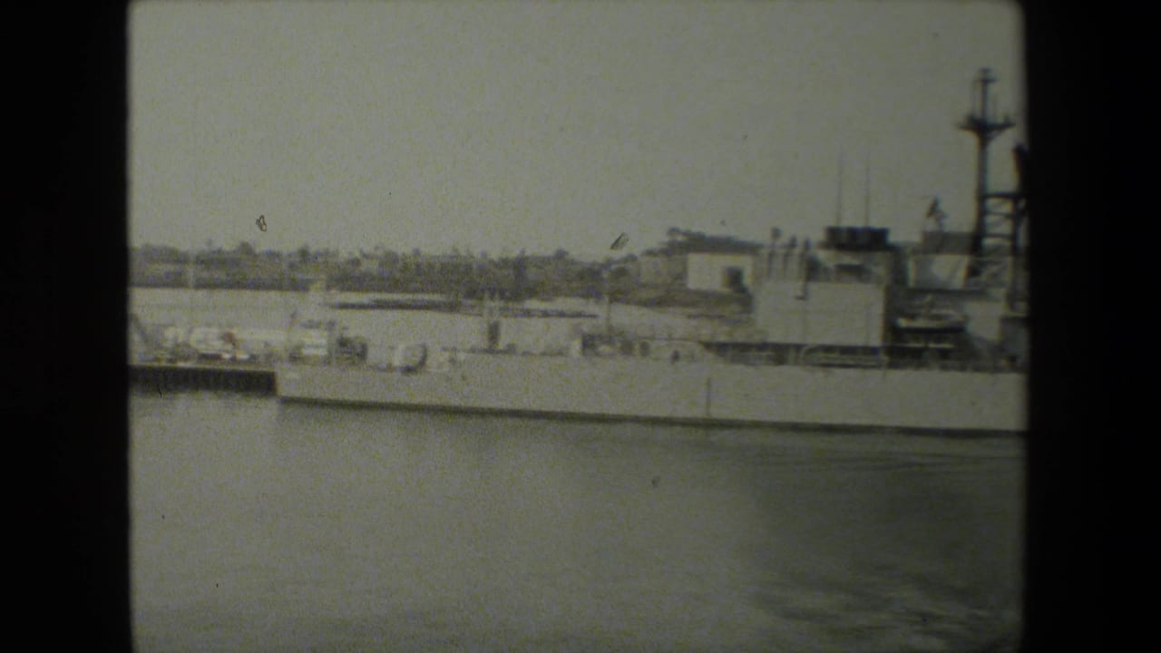The camera continues across the docked naval ship, showing its deck line, superstructure, masts, and harbor water in a soft black-and-white home-movie frame.