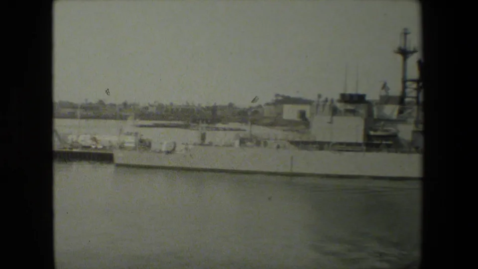 The camera continues across the docked naval ship, showing its deck line, superstructure, masts, and harbor water in a soft black-and-white home-movie frame.