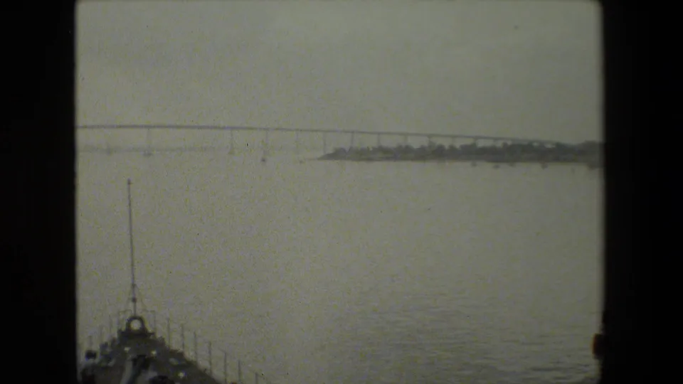 The view shifts away from the ship to open harbor water, a distant low bridge or causeway, shoreline shapes, and the bow or railing of the filming vessel in the foreground.