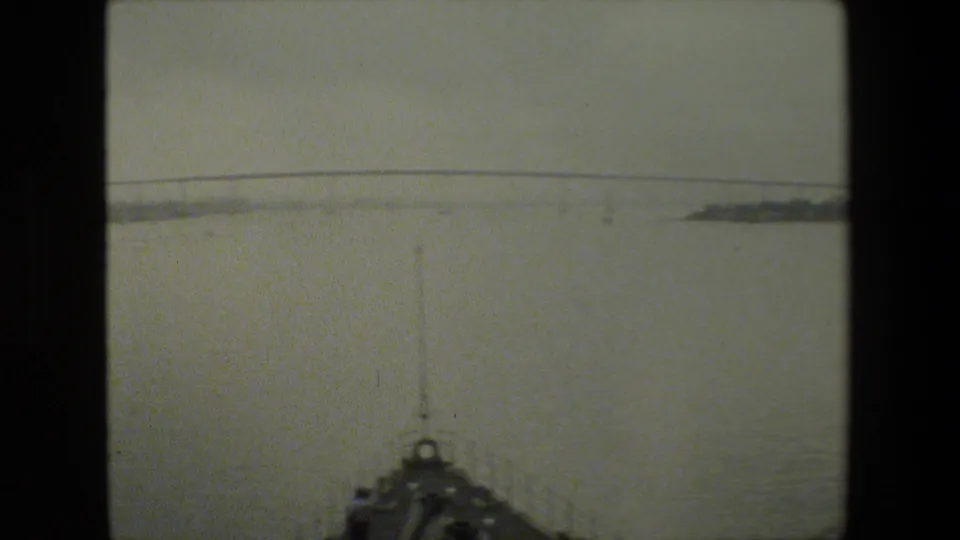 A calm harbor view shows the front of the filming vessel at the bottom of the frame, with distant shoreline and a low bridge or causeway across the water.
