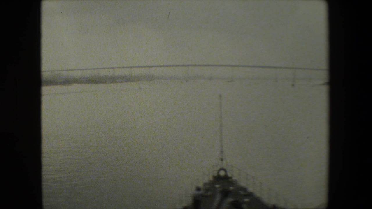 A boat-level 8mm frame shows open water ahead, a distant low bridge across the horizon, and part of the vessel bow at the lower edge.