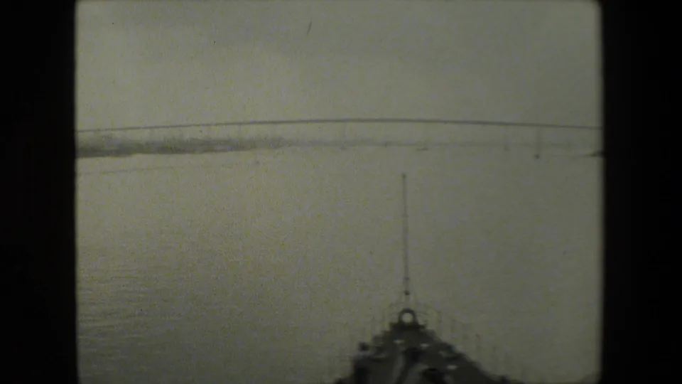 A boat-level 8mm frame shows open water ahead, a distant low bridge across the horizon, and part of the vessel bow at the lower edge.